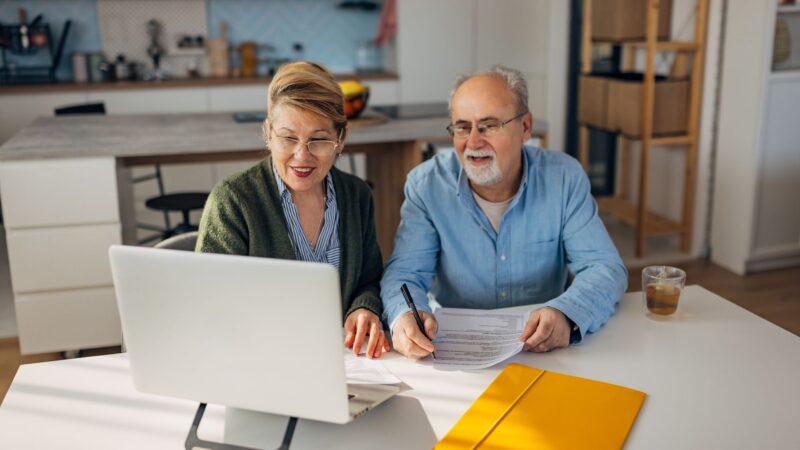 Smiling senior couple manage family budget, sorting out papers, make payments through electronic bank using laptop