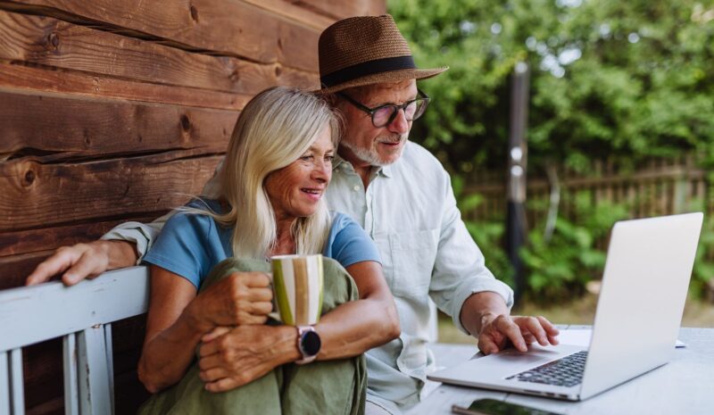 Elderly woman sitting beside her senior husband who is working on laptop.
